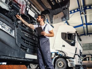 Mechanic using a tablet to inspect a truck engine bay in a trucks and buses service center, with another truck visible in the background